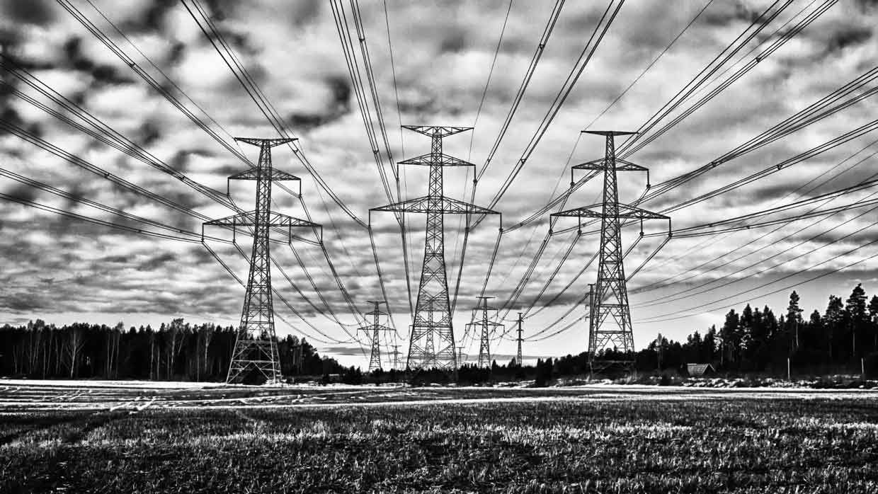 Black-and-white photo of tall electrical transmission towers with power lines stretching across a field under a dramatic cloudy sky.
