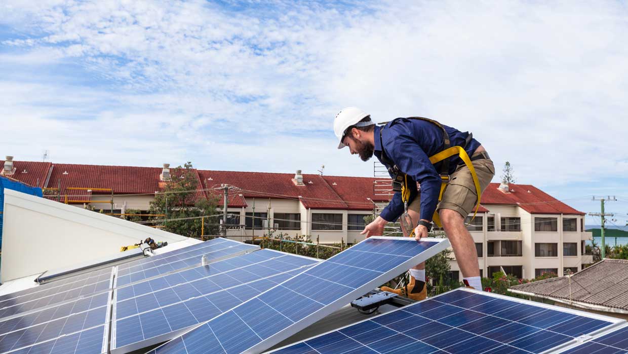 A worker in a hard hat and safety harness installing solar panels on a rooftop, with residential buildings and a partly cloudy sky in the background.